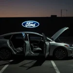 Electric car rolling away from parking space with dim interior visible and Ford logo silhouette in background.