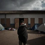 Young migrant stands at abandoned school entrance with boarded windows and tents and hopeful under gray sky
