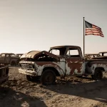Rusty truck sits upright with dusty abandoned lot and faded flag in background.