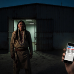 Native American figure standing before abandoned warehouse with glowing smartphone and desert backdrop.