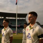 Players stand with black armbands and solemn faces in empty Adelaide Oval cricket pitch with flags half‑staff and muted light