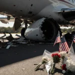 Wreckage lies twisted on ground with broken engine and memorial with flags and flowers beside it