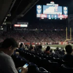 Fans sit on the floor with blank faces and phones while a dim scoreboard shows Titans 20 Chiefs 17.
