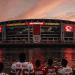 Fans in Chiefs jerseys standing outside Arrowhead Stadium in Kansas City looking up toward a sunset sky with a For Sale sign