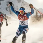 Atle Lie McGrath skiing past Clément Noël, arms raised toward finish line on snowy mountain sunlight