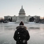 Figure standing at Lady Bird Lake with Texas Capitol in background and snowflakes falling