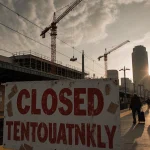 Commuters walking away from boarded-up platform with Closed Temporarily sign and cranes in background.