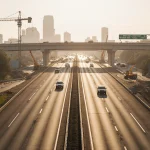 Cars cruising on Austin highway with warm morning light and construction cranes at Westlake & Cedar