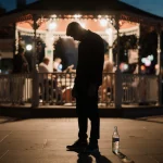 Young man standing with head bowed under dim gazebo and empty bottle against blurred Sixth Street lights