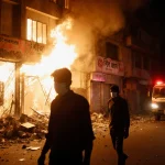 Protesters confront blazing Bangladeshi newspaper office with shattered windows and smoke at night.