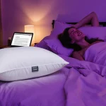 Person lying back on purple bedroom bed with Cloud Pillow and tablet showing article in soft lighting