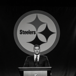Ben Roethlisberger standing at podium with Hall of Honor and stadium lights and blurred Steelers logo in background