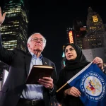 Bernie Sanders and Zohran Mamdani stand with a siddur and flag in NY Times Square beside skyscrapers.