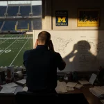 Biff Poggi sits with back to the viewer head in hands staring at a whiteboard with notes in a dim office Michigan memorabilia