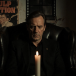 Peter Greene looking down with a flickering candle in a black leather armchair and a nostalgic film reel in the background