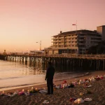 Figure standing on beach sand gazing at sea with candles and flowers reflecting on calm water