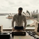 Man sits at cluttered desk with vintage typewriter and briefcase while looking down Bondi Beach backdrop