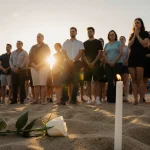 Community mourners standing with a white rose symbolizing remembrance on sand and sunset light over Bondi Beach promenade