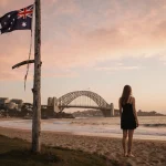 Female figure standing with back to viewer looking out over Bondi Beach at dusk and a torn flagpole