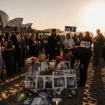 Mourners holding candles and flowers at Bondi Beach with sunset light and the Sydney Opera House in the distance