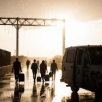 Mexican nationals walking toward a rain‑slick border crossing with a lone open deportation van nearby.