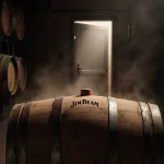 A Jim Beam barrel sits prominently in a misty bourbon barrel room with worn staves and rusty bands.