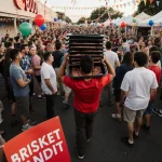 Armando Salazar walking away with stacked brisket trays on his shoulders and red BRISKET BANDIT sign near balloons.