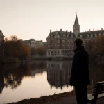 Figure standing near bench looking at river with Brown University buildings reflected and autumn light casting long shadows
