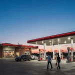 Travelers walking toward Buc‑ee’s travel center at sunrise with white fuel pumps and bright signage