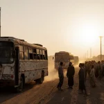 A convoy of rusty buses drives into Jdeidet Yabous with refugees on the outskirts and an American flag fluttering above them.