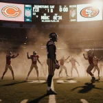 Caleb Williams stands frozen with a slipping football near a glowing 49ers logo on the dusk field while teammates celebrate.