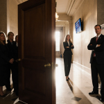 Moderate Republican lawmakers hold hands in Capitol Hill hallway with a large wooden door and a whiteboard marked with red X.
