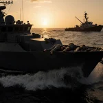 Two lifeless bodies lie on the bow of a black military boat with waves crashing against it and a sunset glow over the Caribbe