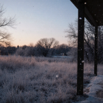 Figure bundled in winter coat standing under porch gazing at snowy landscape with frosted grass and soft dusk light