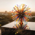 Chihuly glass sculpture standing proudly atop City Arts Center with sunset reflections on its surface and city lights below.