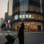 Mother standing in front of a glass birth clinic looking up at dusk with a stroller and a dangling pacifier