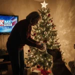 Parent checking Christmas tree water level with a child looking up with holiday cheer while a KXAN news logo glows on the TV