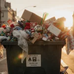 Cluttered return bin overflowing with discarded holiday packaging and broken toys with soft golden sunset light on city stree