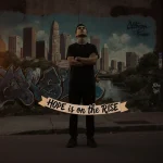 Lone figure standing looking up with graffiti wall and banner reading Hope is on the Rise in dusk light.