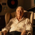 Cliff Richard sits in a plush armchair with vintage microphones and vinyl records in a warm golden light.