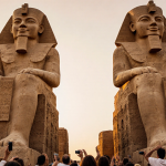 Colossi of Memnon standing with stone feet and golden sunset background and crowd of excited visitors with cameras