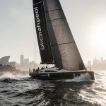 Yacht Comanche sails ahead of LawConnect across Bass Strait with sunrise over Sydney skyline and waves crashing around.