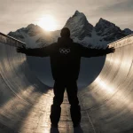 Danny Kass poses at halfpipe base with arms outstretched near snow-capped Italian Alps and sunset glow.