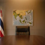 Two empty chairs facing a wooden table in a dim conference room with a Thai flag and map showing diplomatic tension