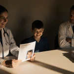 Seated young person listens to two doctors with warm smile and stern posture and prescription pad showing child’s face in lig