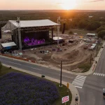 Large concert venue under construction dominates sunset view with wildflowers and protest signs.