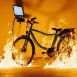 E‑bike charging at station with flames licking its frame and a blurred laptop reflection behind