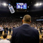 Ed Cooley standing looking at fans in stadium with a water bottle and scoreboard showing 80-77