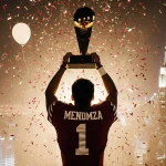 Fernando Mendoza raises arms in triumph with Heisman Trophy at feet confetti around him Indiana jersey against NYC skyline