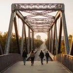 Pedestrians strolling across Cedar Street bridge with warm sunlight glinting on steel arches and green trees in background